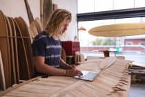 Homem branco fabricante de pranchas de surf em seu estúdio, trabalhando em um projeto usando seu laptop, com pranchas de surf em um rack no fundo . — Fotografia de Stock