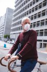 Hombre caucásico mayor por las calles de la ciudad durante el día, con una máscara facial contra el coronavirus, covid 19, montando su bicicleta. - foto de stock
