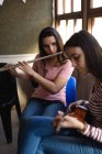 Seitliche Nahaufnahme von zwei kaukasischen Teenager-Mädchen mit langen dunklen Haaren, die vor einem Fenster sitzen und Flöte und Ukulele spielen — Stockfoto