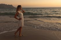 Mulher caucasiana vestindo um vestido branco de pé descalço em uma praia ao pôr do sol, olhando para o mar, relaxando durante um feriado ativo na praia à beira-mar — Fotografia de Stock