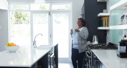 Старша змішана жінка насолоджується своїм часом вдома, соціальною дистанцією та самоізоляцією в карантині, стоїть на її кухні, тримаючи чашку і посміхаючись — стокове фото