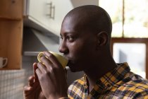 Vista lateral de perto de um homem afro-americano em casa, de pé na cozinha bebendo uma xícara de café e olhando para longe — Fotografia de Stock