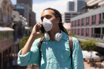 Frontansicht eines Mischlingsmenschen mit langen Dreadlocks, der an einem sonnigen Tag in der Stadt unterwegs ist, mit einer Coronavirus-Maske auf der Straße steht und ein Smartphone benutzt — Stockfoto