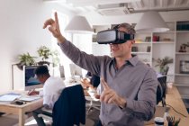 Ein kaukasischer Geschäftsmann arbeitet in einem modernen Büro, trägt ein VR-Headset und berührt einen virtuellen interaktiven Bildschirm, während seine Geschäftspartner im Hintergrund an Schreibtischen arbeiten — Stockfoto