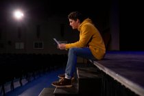 Вид сбоку на кавказского подростка, сидящего на краю сцены с сценарием в пустом школьном театре во время репетиций спектакля — стоковое фото