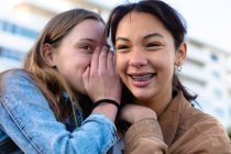 Nahaufnahme einer kaukasischen und einer gemischten Rasse Mädchen genießen die Zeit zusammen hängen an einem sonnigen Tag, lächelnd und flüsternd, Mädchen mit Zahnspange. — Stockfoto
