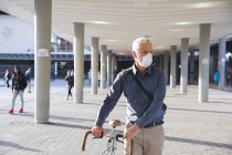 Uomo anziano caucasico in giro per le strade della città durante il giorno, indossando una maschera contro il coronavirus, covid 19, ruote una bicicletta. — Foto stock