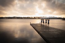 Vue arrière d'une équipe d'aviron de quatre femmes caucasiennes portant un bateau sur leurs épaules, debout sur une jetée dans la rivière au lever du soleil — Photo de stock