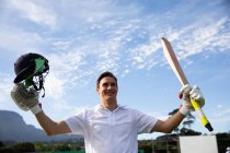 Vorderansicht eines jugendlichen kaukasischen männlichen Cricketspielers, der Weiße trägt, auf dem Spielfeld steht, lächelt und die Hände hebt, einen Cricketschläger und einen Crickethelm in der Hand hält. — Stockfoto