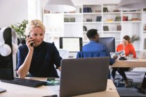 Uma mulher de negócios caucasiana sorridente trabalhando em um escritório moderno, sentada em uma mesa e usando um computador, falando em um smartphone, com seus colegas de negócios trabalhando em segundo plano — Fotografia de Stock