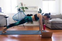 Caucasian woman spending time at home, wearing sportswear, doing a plank, joining online yoga course, using her laptop. Social distancing and self isolation in quarantine lockdown. — Stock Photo