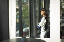 An Asian businesswoman working in a modern office, looking through a window and thinking, crossing her arms, on a sunny day — Stock Photo