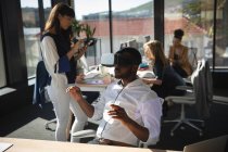 Um empresário afro-americano trabalhando em um escritório moderno, vestindo fone de ouvido VR, tocando tela interativa virtual, com seus colegas trabalhando em segundo plano — Fotografia de Stock