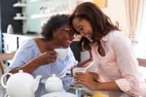 Старшая женщина смешанной расы проводит время дома со своей дочерью, социальное дистанцирование и самоизоляция в карантинной изоляции, пьет чай вместе и разговаривает — стоковое фото