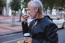 Homme caucasien âgé, portant des vêtements décontractés, dans les rues de la ville pendant la journée, en utilisant un smartphone et tenant une tasse de café à emporter. — Photo de stock