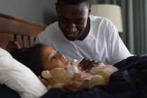 Hombre afroamericano despertando a su hija, durmiendo en su cama y abrazando a un oso de peluche, durante el distanciamiento social en casa durante el bloqueo de cuarentena. - foto de stock