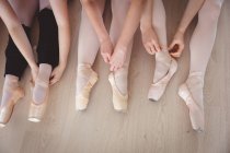Baixa seção de um grupo de dançarinas de balé do sexo feminino amarrando seus sapatos de balé em um estúdio de balé brilhante, se preparando para uma aula de balé, sentado no chão . — Fotografia de Stock