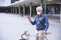 Старший кавказский мужчина днем бродит по улицам города в маске против коронавируса, ковид 19, катает велосипед и пользуется смартфоном. — стоковое фото