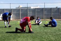 Multi étnico masculino cinco jogadores de futebol um lado vestindo um treinamento de strip equipe em um campo de esportes ao sol, aquecendo-se amarrar seus atacadores. — Fotografia de Stock