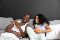 Vista frontale di un uomo afroamericano e della sua giovane figlia seduti su un divano insieme al mattino nel loro soggiorno con ciotole di cereali — Foto stock