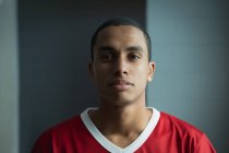 Portrait of a teenage mixed race male field hockey player, wearing a red team strip, standing in a changing room looking straight to camera, focusing before a game — Stock Photo