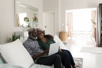 Ein älteres afroamerikanisches Paar, das während der Coronavirus-Epidemie 19 Zeit zu Hause verbringt, soziale Distanzierung und Selbstisolierung in Quarantäne, sitzt auf einem Sofa, umarmt sich, benutzt einen Laptop — Stockfoto
