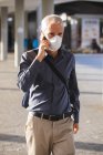 Homem caucasiano sênior para fora e aproximadamente nas ruas da cidade durante o dia,, usando uma máscara facial contra coronavírus, covid 19, usando um smartphone . — Fotografia de Stock