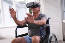 A retired senior Caucasian man at home, sitting in a wheelchair in his underwear in front of a window on a sunny day using a VR headset with his arms outstretched in front of him, self isolating during coronavirus covid19 pandemic — Stock Photo