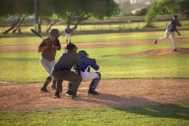 Вид сзади на белого бейсболиста во время игры в бейсбол в солнечный день, готовящегося ударить по мячу бейсбольной битой, кэтчер и другой игрок сидят на корточках за киллером — стоковое фото
