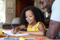 Afroamerikanerin in gelber Bluse, soziale Distanzierung zu Hause während der Quarantäne, am Tisch sitzend und Bilder mit ihrem Vater zeichnend. — Stockfoto