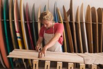 Homem caucasiano fabricante de pranchas de surf trabalhando em seu estúdio, cortando listras de madeira e se preparando para fazer uma prancha de surf, com pranchas de surf em um rack no fundo — Fotografia de Stock