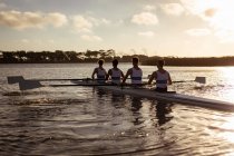 Rear view of a rowing team of four Caucasian men training and rowing on the river, sitting in a rowing boat at sunset — Stock Photo