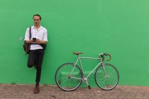 Вид спереди на смешанного гонщика с длинными дредами в городе в солнечный день, стоящего на улице, прислонившись к зеленой стене, используя смартфон, с велосипедом, стоящим рядом с ним. — стоковое фото