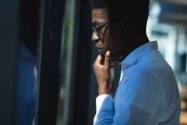 Un homme d'affaires afro-américain aux cheveux courts, portant une chemise blanche et des lunettes debout à l'intérieur d'un café et regardant la fenêtre toucher son menton — Photo de stock
