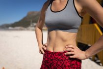Vue de face section médiane de la femme, vêtue de vêtements de sport, debout avec les mains sur les hanches sur la plage, relaxant après le jogging. . — Photo de stock