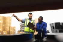An African American male worker and a Caucasian male supervisor in a storage warehouse at a factory making wheelchairs, standing and talking, holding a clipboard — Stock Photo