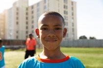 Портрет змішаної раси хлопчика-футболіста в синій командній смузі, що стоїть на ігровому полі в сонячний день, дивлячись на камеру і посміхаючись, з товаришами по команді, що стоять на задньому плані — стокове фото