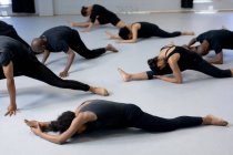 Vue latérale d'un groupe multi-ethnique de danseurs modernes, hommes et femmes, en tenue noire, pratiquant une routine de danse lors d'un cours de danse dans un studio lumineux, allongés sur le sol et s'étirant vers le haut. — Photo de stock