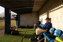 Вид сбоку на двух бейсболистов смешанной расы, во время игры в бейсбол в солнечный день, сидящих на скамейке, со своим товарищем по команде, болеющим за свою команду, готовящимся выйти на поле — стоковое фото