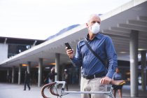 Älterer kaukasischer Mann, der tagsüber auf den Straßen der Stadt unterwegs ist, Gesichtsmaske gegen Coronavirus trägt, 19 Jahre alt ist, Fahrrad fährt und ein Smartphone benutzt. — Stockfoto