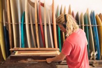 Homem caucasiano fabricante de pranchas de surf trabalhando em seu estúdio, usando fones de ouvido de proteção, cortando listras de madeira e se preparando para fazer uma prancha de surf, com pranchas de surf em um rack no fundo . — Fotografia de Stock