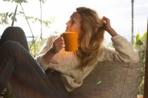 Vista laterale di una donna caucasica seduta nel suo salotto di fronte a una finestra in una giornata di sole, con una tazza e un sorriso — Foto stock