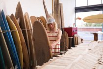 Caucasian male surfboard maker in his studio, inspecting one of the surfboards, with other surfboards in a rack behind him. — Stock Photo