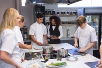 Caucasian male chef cutting vegetables, talking to mixed race waitress, with other chefs cooking in the foreground. Cookery class at a restaurant kitchen. — Stock Photo