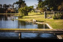Un molo in un lago su un parco e alberi su una riva erbosa con recinzioni in una giornata di sole — Foto stock