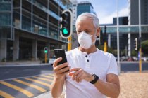 Älterer Kaukasier, der tagsüber auf den Straßen der Stadt unterwegs ist und eine Gesichtsmaske gegen Coronavirus trägt, 19 Jahre alt, Smartphone benutzt. — Stockfoto