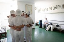 Side view of a group of teenage multi-ethnic male cricket players wearing whites, huddling in a changing room, with another player resting on a bench. — Stock Photo