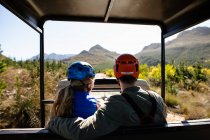 Rückansicht eines kaukasischen Paares, das an einem sonnigen Tag in den Bergen in Reißverschlussausrüstung im Auto sitzt und die Zeit in der Natur genießt — Stockfoto