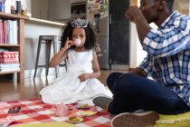 Chica afroamericana con una corona de juguete, distanciamiento social en casa durante el bloqueo de cuarentena, jugando con su padre, teniendo una fiesta de té de muñecas. - foto de stock