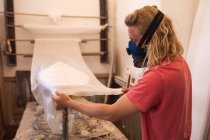 Homem caucasiano fabricante de pranchas de surf vestindo uma máscara facial de respiração, trabalhando em seu estúdio, inspecionando uma prancha de surf coberta com um pedaço de pano branco . — Fotografia de Stock